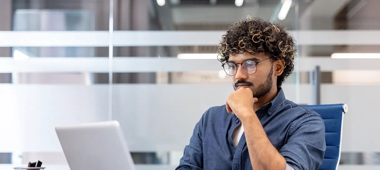 An advertiser sits at a laptop in the office, looking at the screen and thinking.